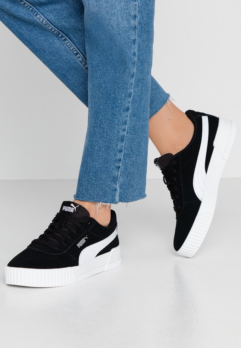 Černé semišové tenisky s bílými akcenty, plochými tkaničkami a silnou gumovou podrážkou. Úzký design s logem na jazyku a na boku.