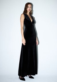 Zwarte fluwelen avondjurk met een diepe V-hals, mouwloos ontwerp en een aansluitende silhouet, die tot op de grond valt. Zwarte spitse hakken.