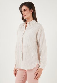 Light beige button-up shirt with a textured weave, featuring long sleeves, a classic collar, and a curved hemline.