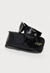 Ochelari de soare cu rame negre lucioase și lentile întunecate, așezați pe un toc de piele texturată neagră, cu accente aurii ale logo-ului.