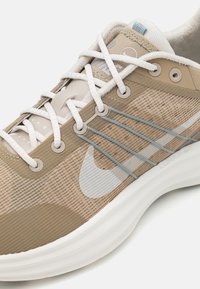 Scarpa sportiva beige con una tomaia texture, dettagli grigi e lacci bianchi. Presenta una suola arrotondata con il marchio "LUNAR".