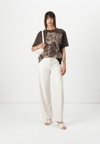 Braunes Grafik-T-Shirt mit stilisierten Tierdesigns, kombiniert mit weißen weit geschnittenen Hosen und cremefarbenen offenen Schuhen mit einem Detail.