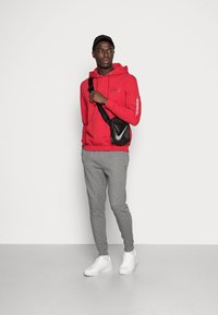Felpa rossa con cappuccio e un logo sul petto, con testo sulla manica, abbinata a pantaloni della tuta grigi e sneakers bianche, completata da una borsa a tracolla nera.