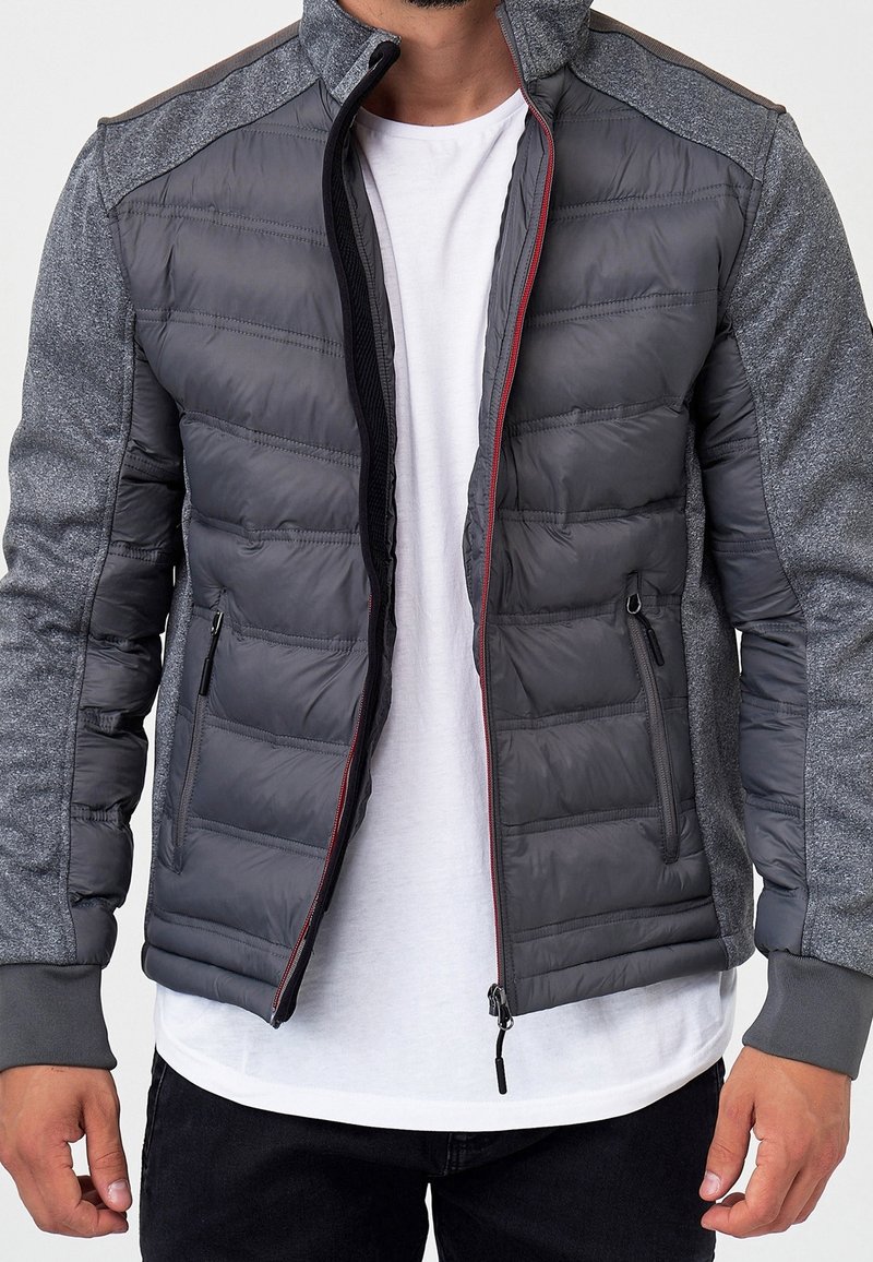 Mann trägt eine teilweise geschlossene graue gesteppte Jacke mit Strickärmeln über einem weißen T-Shirt und schwarzen Hosen, steht vor einem schlichten Hintergrund.