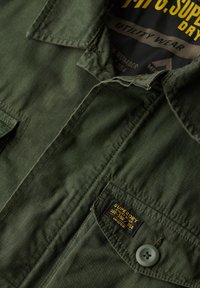 Camisa utilitaria verde oliva con cuello abotonado, dos bolsillos frontales y una etiqueta con texto en amarillo y negro. Tejido texturizado con líneas visibles.