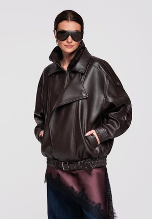 Kvinde iført oversized mørkebrun læderjakke, store sorte solbriller, guld øreringe i hoop-stil og mørkelilla satin nederdel med sort blonderkant.