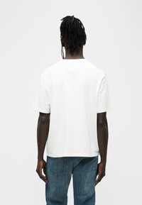 Λευκό βαμβακερό μπλουζάκι με κοντά μανίκια, στρογγυλή λαιμόκοψη και χαλαρή εφαρμογή. Φορεμένο με μπλε τζιν παντελόνι, φαίνεται από πίσω.