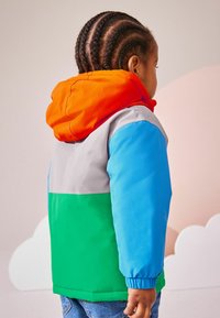 Chaqueta con bloques de color que presenta una capucha naranja, parte superior gris, mangas azules y sección inferior verde, fabricada con material ligero y resistente al agua.