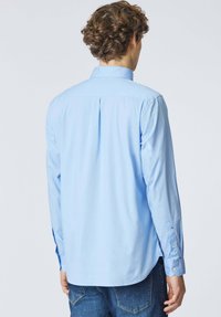 Uomo con capelli castani ricci che indossa una camicia azzurra a maniche lunghe con bottoni e jeans di denim blu, di spalle rispetto alla fotocamera.