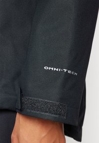 Black jacket close-up with a textured fabric, featuring a velcro strap at the cuff and a white logo reading "OMNI-TECH."