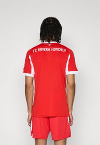 Maillot rouge du FC Bayern München avec des accents blancs, à manches courtes et col ras du cou. Texte au dos indique "FC BAYERN MÜNCHEN."