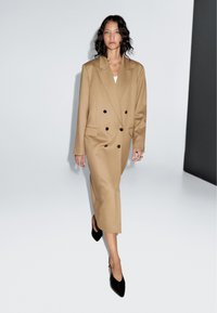 Double-breasted, camel-colored coat dress in a structured design with black buttons. Model wears black slingback heels, minimal accessories.