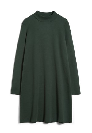 Mujer con vestido oscuro de manga larga hasta la rodilla y botas negras con tacón hasta la mitad de la pantorrilla sobre un fondo claro y liso.
