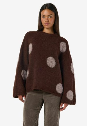 Donna che indossa un ampio maglione marrone scuro con grandi pois grigi chiari e pantaloni in corduroy grigi, in piedi contro uno sfondo bianco.