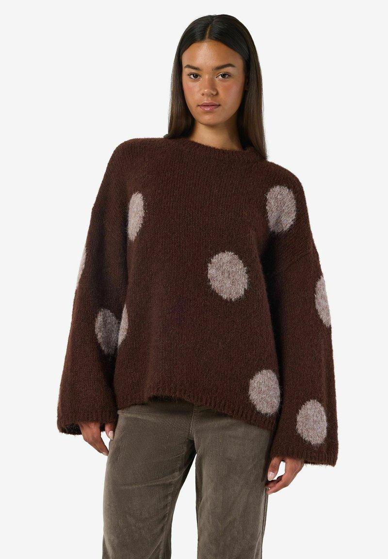 Kvinna som bär en lös mörkbrun tröja med stora ljusgråa prickar och grå kordflorbyxor, stående mot vit bakgrund.