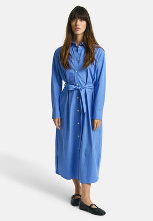 Abito camicia azzurro chiaro a maniche lunghe con bottoni frontali e vita da annodare, caratterizzato da una linea dritta e spacchi laterali, indossato con ballerine nere.