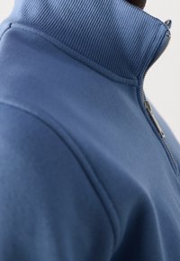Gros plan d'une personne portant un pull bleu à fermeture éclair avec un col montant côtelé sur un fond uni.