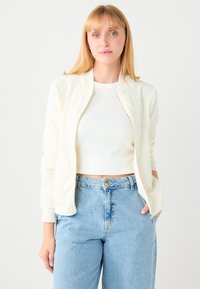 Roomwitte bomberjas met ritssluiting, gestructureerde stof, geribbelde manchetten en bloemrijke borduurwerk. Gecombineerd met een high-waist blauwe jeans en een wit topje.