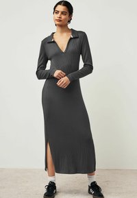 Robe maxi grise côtelée avec des manches longues, un décolleté en V et un col. Présente des fentes sur les côtés et une silhouette ajustée. Portée avec des baskets noires.