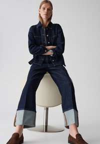 Veste en denim foncé avec des boutons et des poches, associée à un pantalon en denim foncé à large jambe avec des revers bleu clair et des mocassins marron.