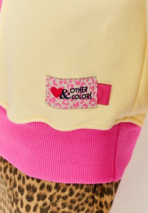 Kolorowy sweatshirt z żółtym korpusem i różowymi ściągaczami, z metką w króliczy wzór, na której widnieje napis "Inne i Kolory" w czarnym kolorze.