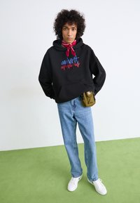Felpa nera con testo colorato, jeans azzurri chiari, sneakers bianche e una piccola borsa verde oliva. Tessuto morbido, vestibilità rilassata, look informale.