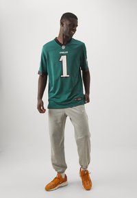 Teal NFL-trøje med grå accenter og stort hvidt "1" nummer, parret med lys grå sweatpants og orange sneakers. Minimalistisk baggrund.