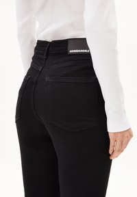 Los jeans negros de talle alto cuentan con bolsillos traseros, costuras estructuradas y una etiqueta de cuero negro con marca en la cintura. La tela tiene una textura suave.