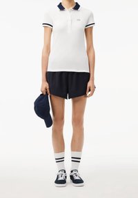 Vit pikétröja med marinblå krage, svarta träningsshorts, marinblå keps i handen, vita strumpor med marinblå ränder och marinblå sneakers.