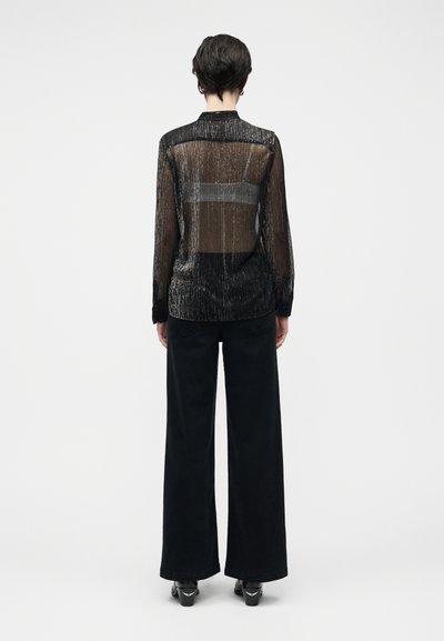 Črna prosojna bluza z navpičnimi črtami, dolgimi rokavi in rahlo sproščenim krojem, kombinirana z visokim pasom in črnimi hlačami širokih hlačnic.