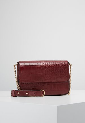 Bolso bandolera de piel burdeos con relieve de cocodrilo, con cadena dorada y sección de piel ajustable, sobre una superficie blanca.