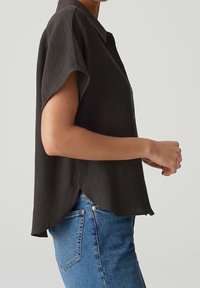Chemise noire à manches courtes avec un tissu texturé, ourlet légèrement incurvé et col, associée à un pantalon en denim bleu.