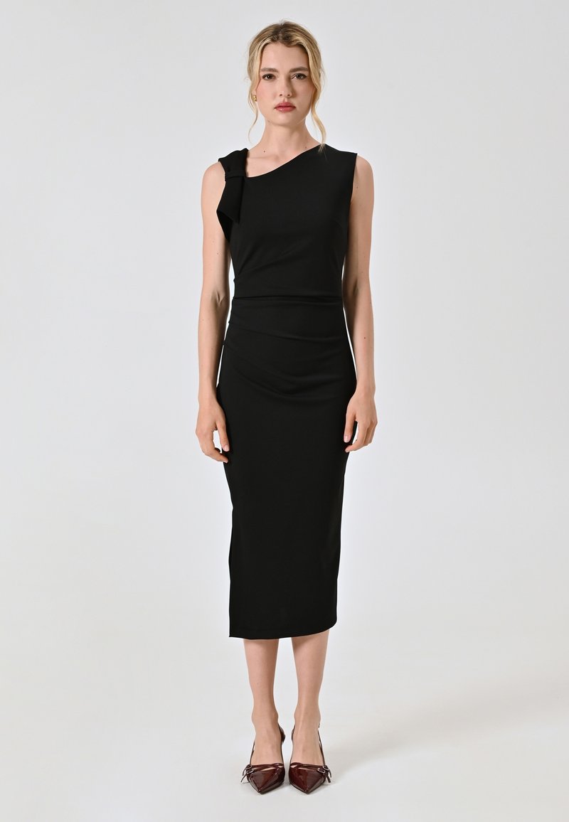 Schwarzes Midi-Kleid mit asymmetrischem Ausschnitt und Rüschendetails. Glatter, enganliegender Stoff mit schlanker Silhouette, kombiniert mit spitzen bordeauxfarbenen Absätzen.