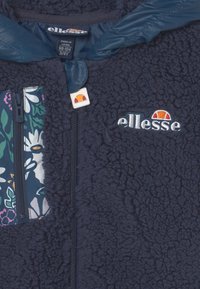 Jaqueta de fleece azul-marinho com um bolso floral com fecho, logótipo bordado e uma forragem de tecido impermeável na gola e na área do fecho.