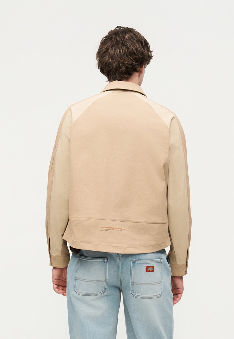 Mann mit braunen Haaren, der eine beige Jacke und hellblaue Jeans trägt, von hinten vor weißem Hintergrund gezeigt.