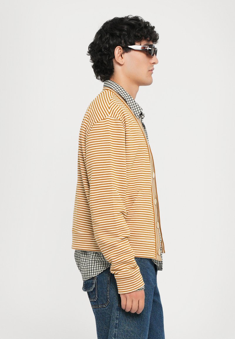 Gestreifter Cardigan in Orange und Gelb, mit Knöpfen, langärmlig. Kombiniert mit einem schwarz-weißen karierten Hemd und blauen Jeans.