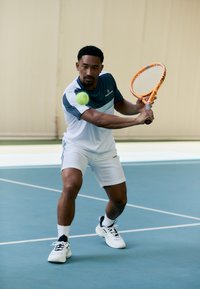 Tennisspeler in een witte en blauwe sportoutfit, met een oranje racket, bereid om een groene tennisbal te slaan op een blauwe tennisbaan.