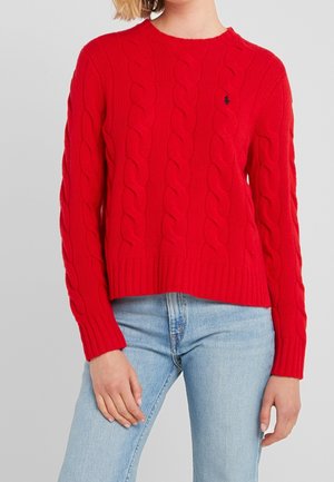 Kvinde iført rød kabelstrikket sweater og lyseblå jeans, stående med en hånd hvilende på låret mod en ensfarvet grå baggrund.