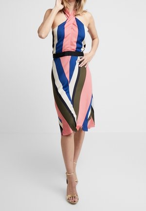 Vrouw draagt een mouwloze halterjurk met roze, blauwe, witte en olijfgroene verticale strepen, gecombineerd met gouden sandalen met enkelbandje.