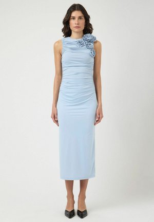 Žena u svijetloplavoj midi haljini bez rukava s tri cvjetna aplikacija na lijevom ramenu, u kombinaciji s crnim cipelama šiljastih vrhova.