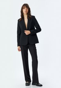 Zwart op maat gemaakt pak met een diepe V-hals blazer, slim-fit broek en gestructureerde zwarte instappers met gouden accenten.