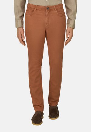 Mand iført slim-fit brændt orange bukser, beige skjorte stoppet ind og brune ruskindsløfler, stående foran en ensfarvet baggrund.