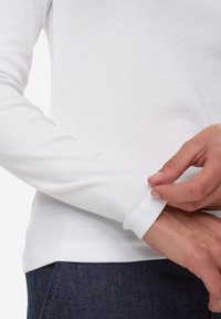 Langärmliges weißes Oberteil aus geripptem Stoff, mit eng anliegenden Bündchen und abgerundetem Saum, kombiniert mit dunklen Jeans.