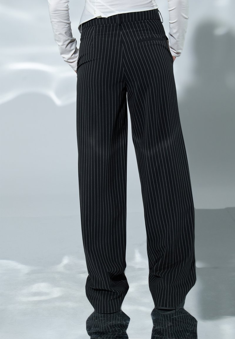 Personne portant un pantalon large noir à fines rayures et un haut blanc à manches longues, debout devant un fond neutre.
