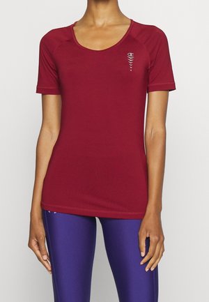 Vrouw draagt een nauwsluitend bordeauxkleurig sportshirt met korte mouwen met een klein logo op de borst en paarse legging tegen een effen achtergrond.