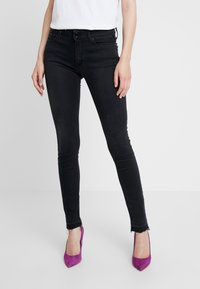 Jeans negros ajustados con una cintura de altura media, dobladillo ligeramente deshilachado y bolsillos frontales. Combinados con zapatos de tacón altos morados.