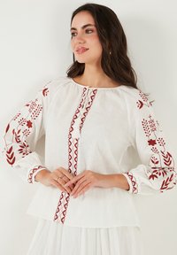 Blouse en lin blanc ornée d'une broderie florale rouge le long des manches et au centre, avec un col rond et des manches bouffantes froncées.