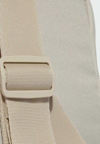 Correa de tela beige con superficie texturizada y un hebilla de plástico. La correa está unida a un bolso a juego, que muestra un material suave y duradero.