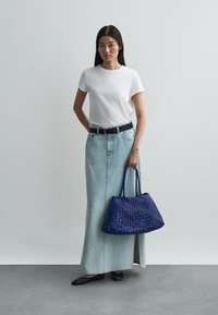 Λευκό T-shirt, ελαφρώς μπλε τζιν μάξι φούστα, μαύρη ζώνη και μαύρες επίπεδες μποέμ. Κρατώντας μια μεγάλη πλεξούδα μπλε τσάντα. Ουδέτερο φόντο.