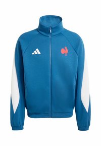 Modrá športová bunda s vysokým golierom, bielymi akcentmi na rukávoch, logom červeného kohúta, zipsovým uzáverom a rebrovanými manžetami a dolným okrajom.